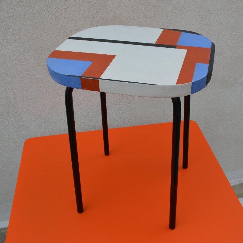 Vue d'ensemble du tabouret Géo Ses pieds sont en acier laqué noir et l'assise d'une épaisseur de 38 mm est recouverte d'un béton ciré blanc muni de motifs en béton ciré bleu, orange et anthracite