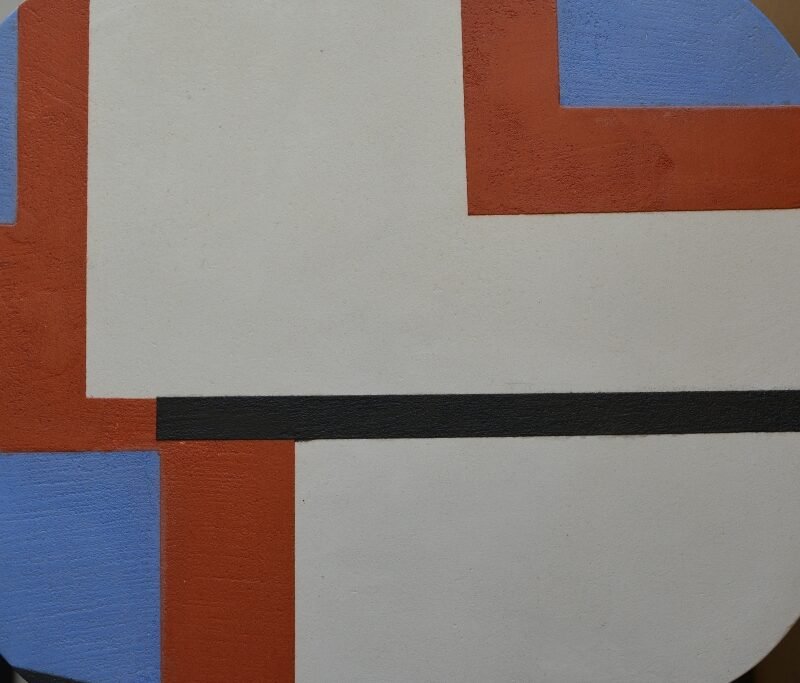 Gros plan sur l'assise d'un tabouret en béton ciré fond blanc avec décoration géométriques bleu, gris anthracite et orange